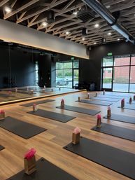 Bright modern yoga studio with wood floors, exposed beam ceiling and mirrored wall; rows of black mats by floor-to-ceiling windows, each paired with a small brown gift bag topped with pink tissue—class-ready.