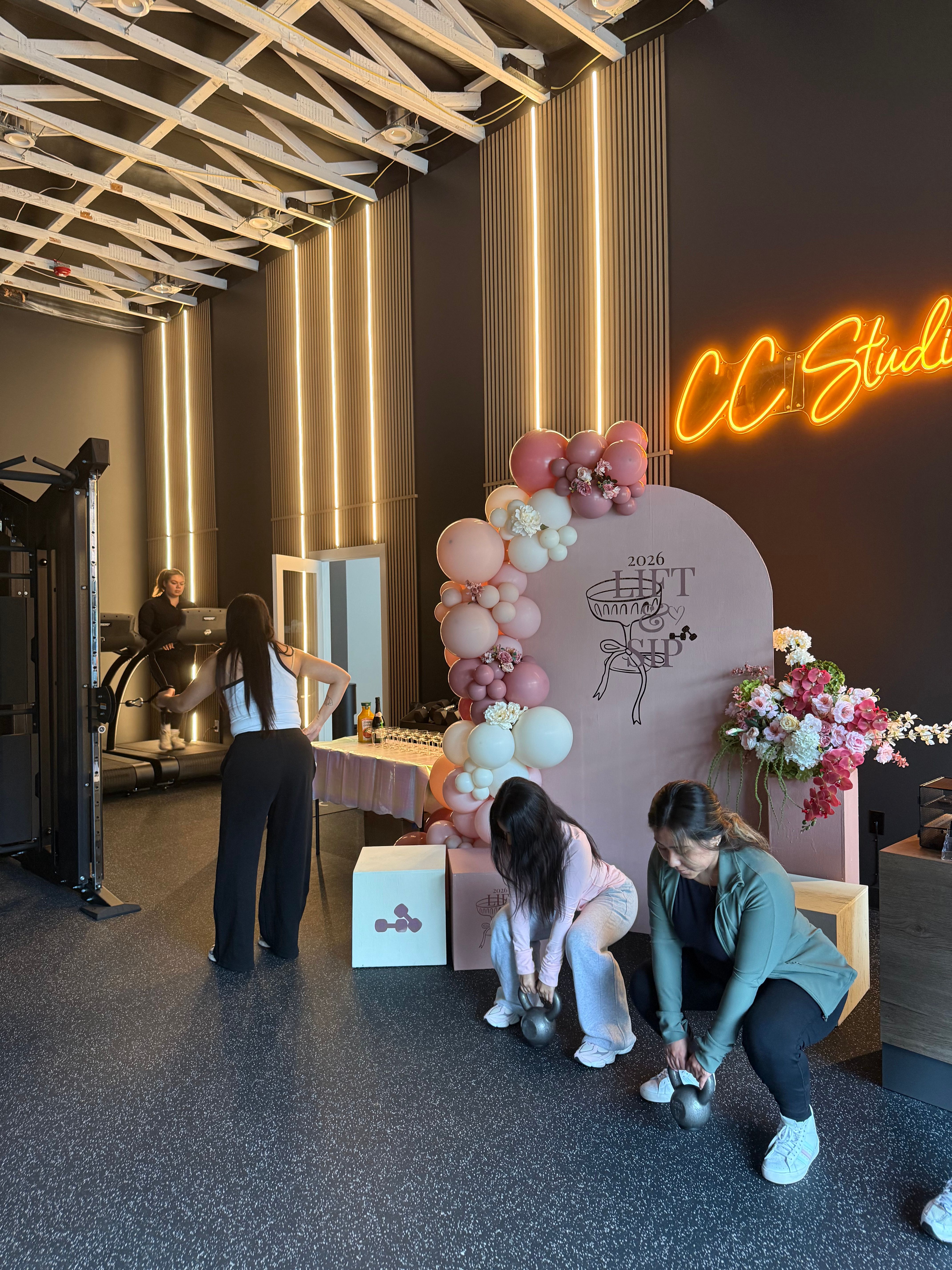 Women exercising in a modern fitness studio decorated for a pink-themed event — kettlebell squats, a treadmill, balloon-and-flower arch backdrop, and a glowing neon studio sign.