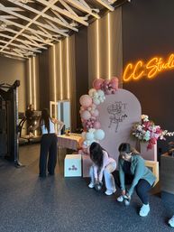Women exercising in a modern fitness studio decorated for a pink-themed event — kettlebell squats, a treadmill, balloon-and-flower arch backdrop, and a glowing neon studio sign.