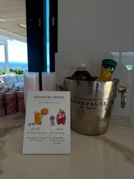 Coastal drink station with a signature drinks menu, stacked plastic cups, sparkling water cans and an ice-filled champagne bucket holding juice bottles on a marble counter with patio and ocean visible through glass doors.