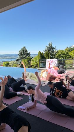 Sunny outdoor group yoga on pink mats beside an infinity pool, overlooking a scenic lake and tree‑covered hills under a clear blue sky.