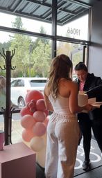 Two people at a glass storefront entrance exchanging a package beside a playful pink balloon column, with parked cars and leafy trees visible outside.