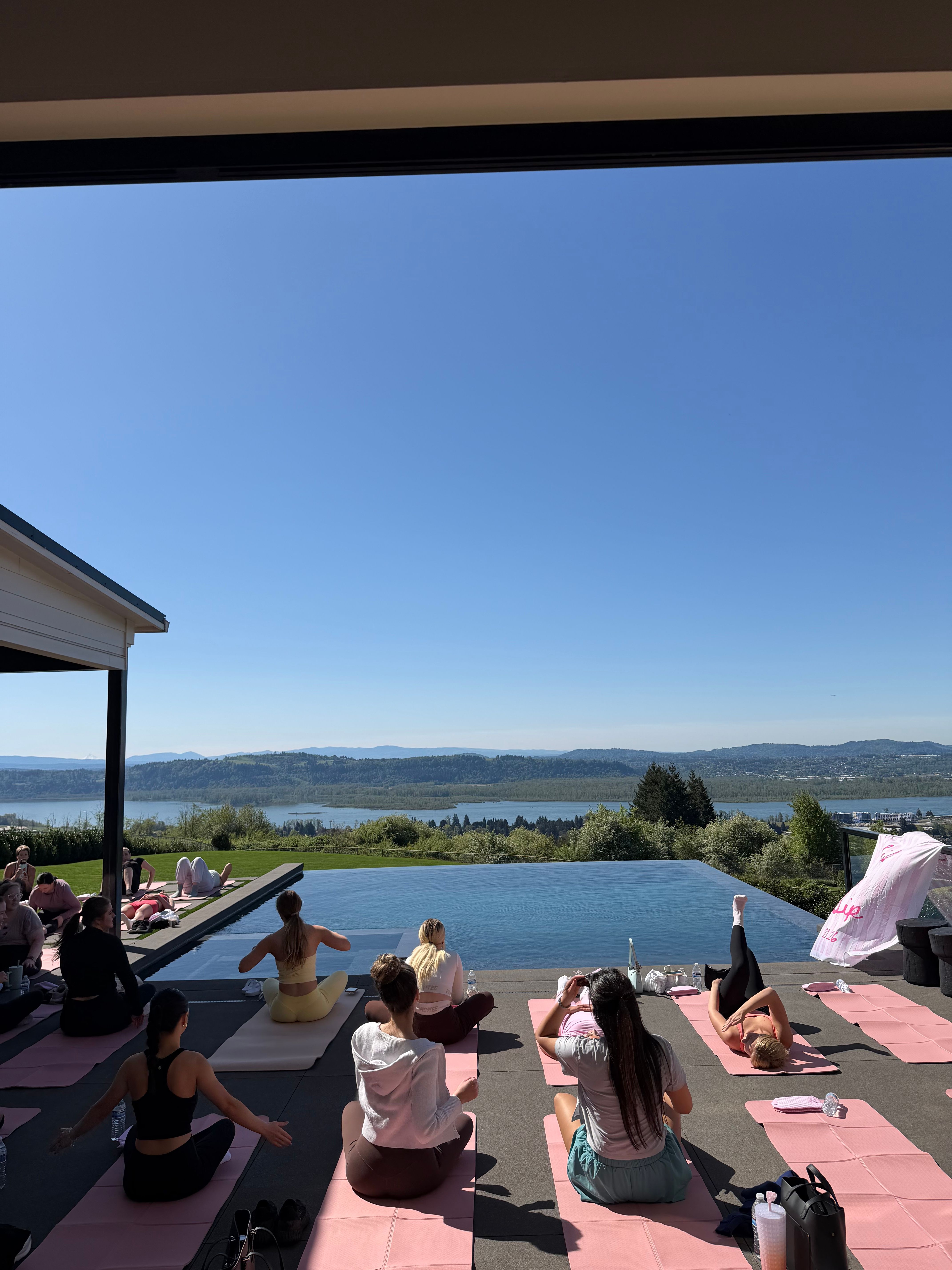 Sunlit outdoor yoga class on pink mats beside an infinity pool, overlooking a river valley and rolling hills under a clear blue sky.
