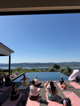 Sunlit outdoor yoga class on pink mats beside an infinity pool, overlooking a river valley and rolling hills under a clear blue sky.