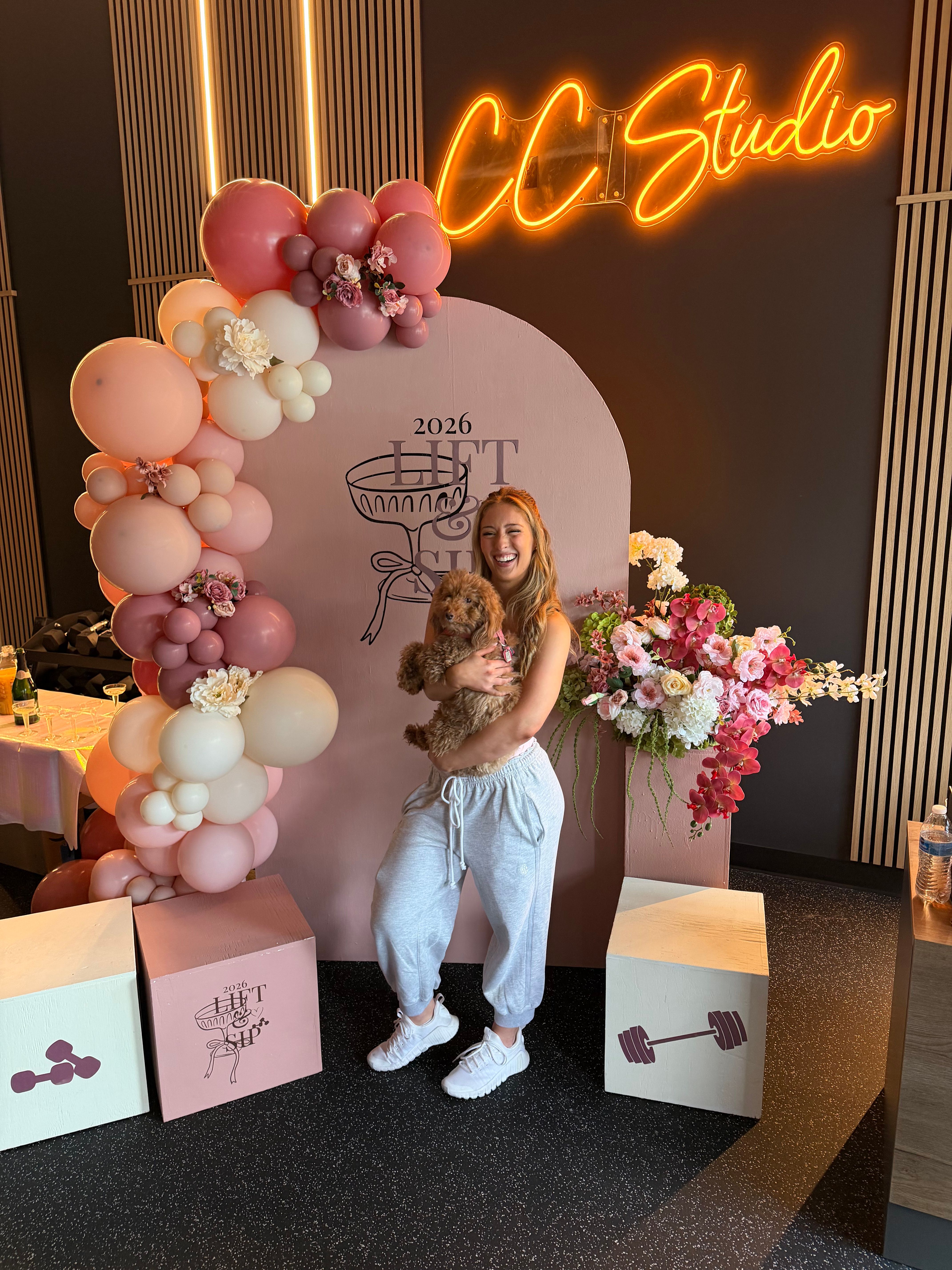 Laughing woman in gray joggers holding a fluffy brown puppy in front of a pink arch decorated with blush balloons and floral arrangements, neon sign glow and wood-accent wall in the background.