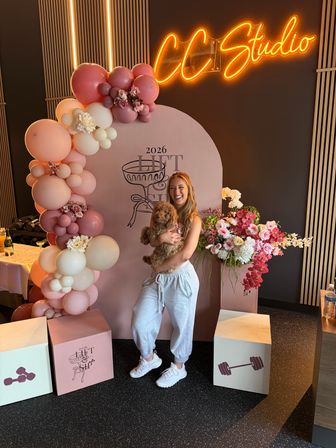 Laughing woman in gray joggers holding a fluffy brown puppy in front of a pink arch decorated with blush balloons and floral arrangements, neon sign glow and wood-accent wall in the background.