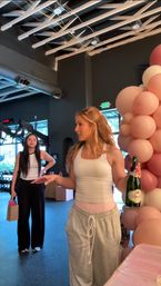 Two women in a modern indoor fitness studio celebrating by a pink balloon garland; one in a white tank and gray sweatpants holds a champagne bottle while gym equipment and industrial ceiling fixtures are visible.