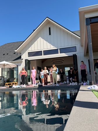 Group of women in colorful activewear doing a poolside fitness/yoga session in front of a modern white farmhouse with open doors and a gabled roof, reflected in the calm blue pool under a clear sky.