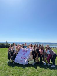Sunny hilltop group photo of about two dozen people on a green lawn overlooking a river valley and distant hills, holding a pink-striped banner reading “Lift & Sip 2026” for an outdoor fitness retreat vibe.