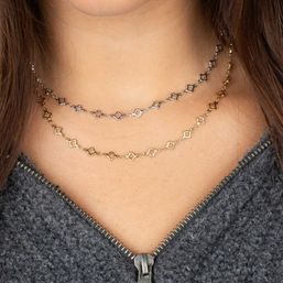Close-up of layered dainty gold and silver geometric choker necklaces with small open quatrefoil links on a neckline above a grey zip-up fleece.