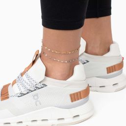 Close-up of ankles wearing stacked gold and silver chain anklets with white athletic sneakers featuring tan accents and black cropped leggings — casual streetwear look.
