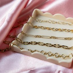 Close-up of assorted gleaming gold chain necklaces arranged on a scalloped white jewelry tray atop soft pink satin fabric