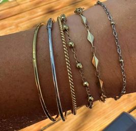 Chic mixed-metal bracelet stack on a wrist — thin gold and silver bangles, twisted rope, beaded chain and geometric link bracelets against a sunlit wooden deck background.