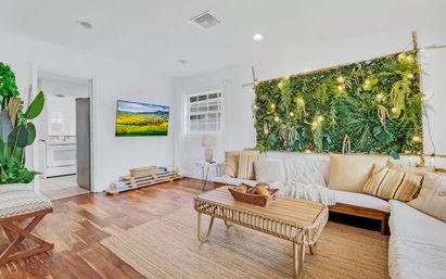 Bright modern living room with a lush vertical plant wall lit by string lights, white sectional sofa with neutral pillows, rattan coffee table, hardwood floors, indoor plants, and an open doorway to a white kitchen.