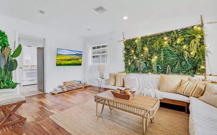 Bright modern living room with a lush vertical plant wall lit by string lights, white sectional sofa with neutral pillows, rattan coffee table, hardwood floors, indoor plants, and an open doorway to a white kitchen.