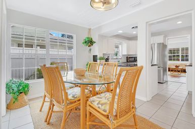 Bright, coastal-style dining area with six rattan chairs around a round glass table, woven rug, hanging plant, large windows and an open white kitchen with stainless steel appliances.