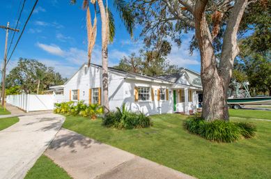 Sunny white coastal bungalow with green front door, palm trees and large oak on a manicured lawn beside a sidewalk and a boat on a trailer
