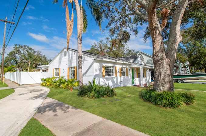 Sunny white coastal bungalow with green front door, palm trees and large oak on a manicured lawn beside a sidewalk and a boat on a trailer