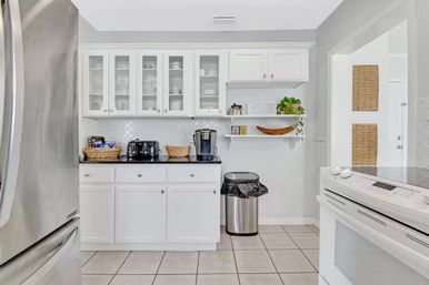 Bright white kitchen with glass-front cabinets, stainless steel refrigerator and appliances, black countertop coffee station, open shelves with plant, and tiled floor