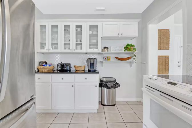 Bright white kitchen with glass-front cabinets, stainless steel refrigerator and appliances, black countertop coffee station, open shelves with plant, and tiled floor