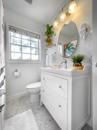 Small modern white bathroom with hexagon tile floor, white vanity and sink, round wall mirror under a three-bulb light, potted plants on the vanity and floating shelf, and a toilet by a window with blinds.