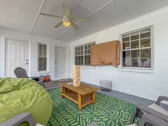 Cozy covered porch with white walls and ceiling fan, green leaf-pattern rug, lime bean bag and gray chairs, wooden coffee table holding a large Jenga tower, and a covered outdoor TV.