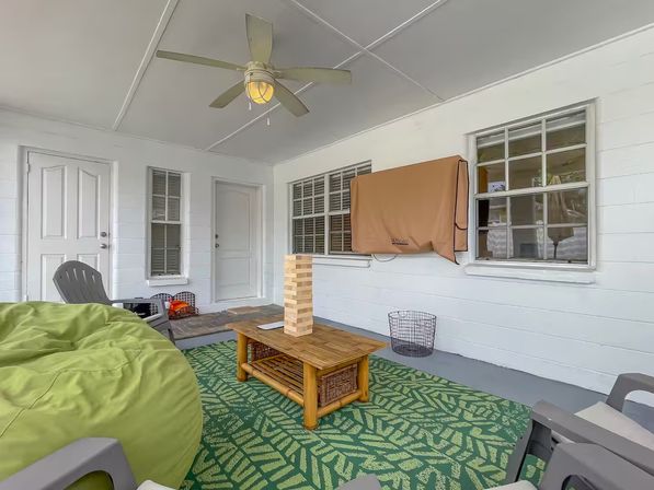 Cozy covered porch with white walls and ceiling fan, green leaf-pattern rug, lime bean bag and gray chairs, wooden coffee table holding a large Jenga tower, and a covered outdoor TV.