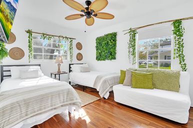 Bright sunlit twin bedroom with white bedding and striped throws, white daybed with green pillows, hanging faux greenery and woven wall decor, tropical-style ceiling fan and polished hardwood floors.