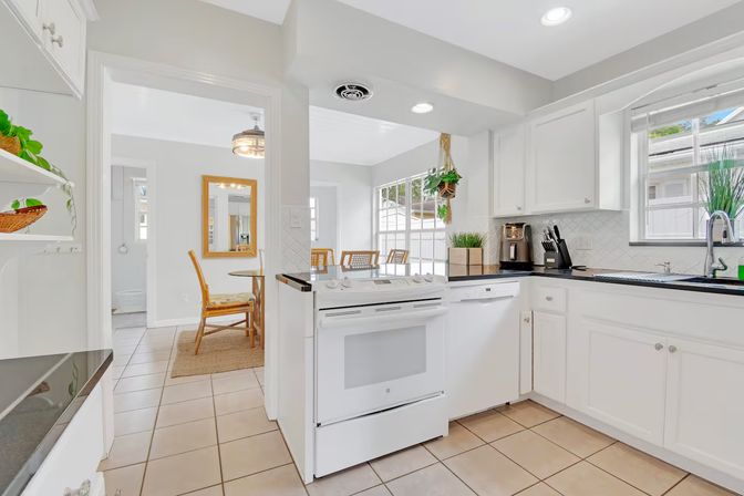 Bright white kitchen with tiled floor, electric stove and dishwasher, black countertops, hanging plant and sink under a window, opening to a sunny dining nook with wooden chairs.