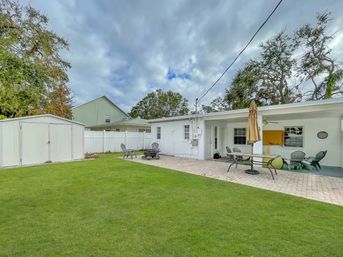 Residential backyard with green lawn, paver patio, white storage shed and fence; single-story white house with covered patio, outdoor dining table and umbrella, Adirondack chairs around a fire pit under an overcast sky.