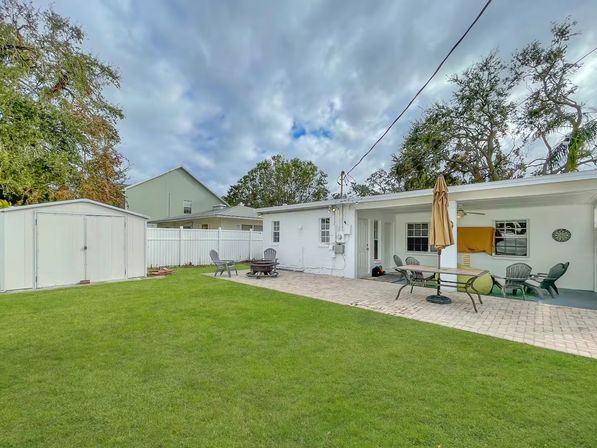 Residential backyard with green lawn, paver patio, white storage shed and fence; single-story white house with covered patio, outdoor dining table and umbrella, Adirondack chairs around a fire pit under an overcast sky.