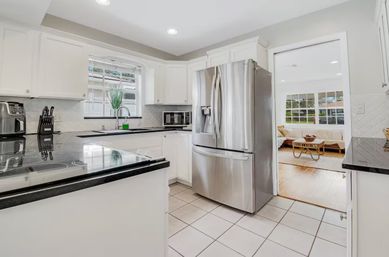 Bright modern white kitchen with black countertops and island cooktop, stainless steel French-door refrigerator, tiled floor, sink under window with potted plant, and an open doorway to a sunlit living room