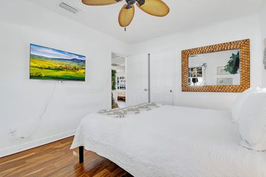 Bright modern bedroom with white quilted bed, warm hardwood floors, leaf-blade ceiling fan, wall-mounted TV showing a green landscape, and a woven-frame mirror.