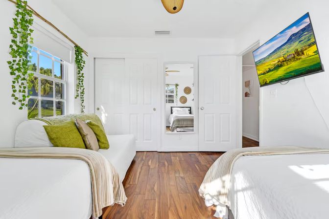 Bright airy bedroom with white walls and warm wood floors, twin beds with green accent pillows, hanging ivy by the window and a wall-mounted TV