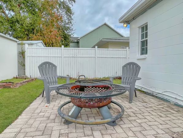 Cozy suburban backyard patio with a circular metal fire pit on brick pavers, two gray Adirondack-style chairs, a white privacy fence and house siding under a cloudy sky.