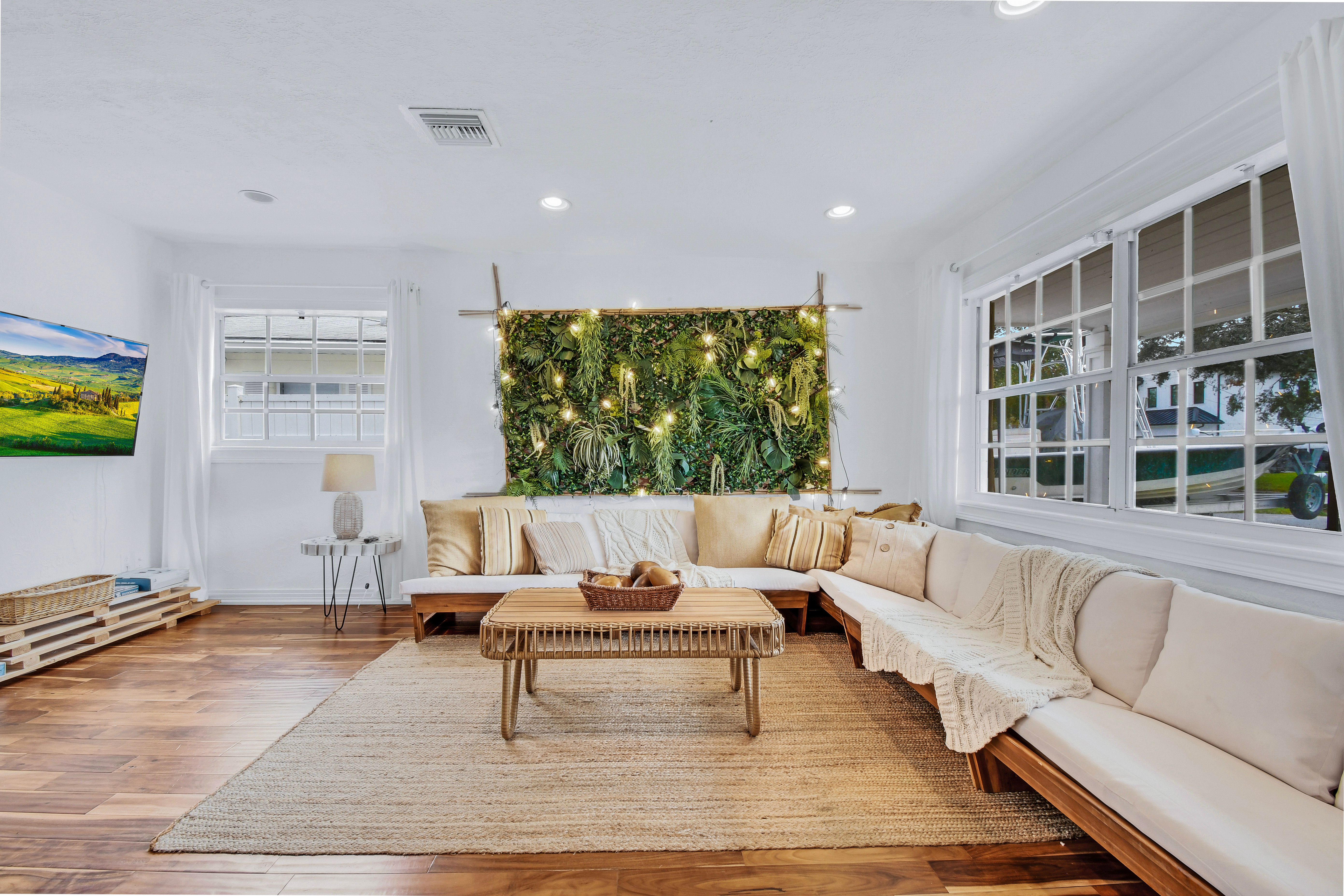 Bright, cozy living room with a large L-shaped beige sectional, woven rattan coffee table, jute rug, illuminated vertical plant wall with string lights, hardwood floors and oversized windows letting in natural light.
