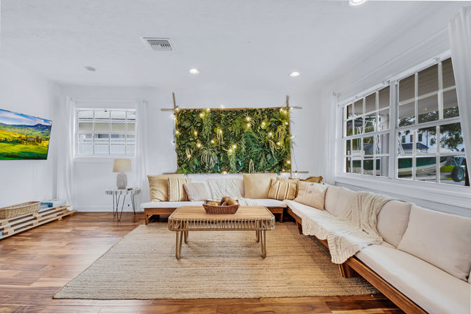 Bright, cozy living room with a large L-shaped beige sectional, woven rattan coffee table, jute rug, illuminated vertical plant wall with string lights, hardwood floors and oversized windows letting in natural light.
