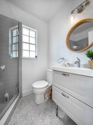 Bright, airy small apartment bathroom with glass walk-in shower, hexagon marble tile floor, white vanity with round wood-framed mirror, compact toilet and frosted window letting in natural light.
