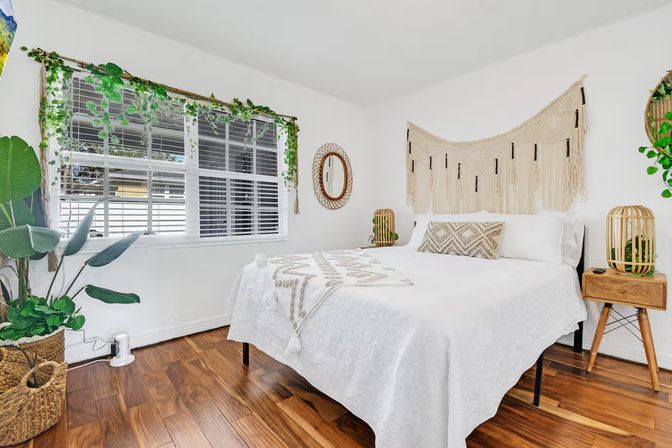 Sunlit boho bedroom with white quilted bed, macramé wall hanging, patterned throw pillow, wooden nightstands with rattan lamps, lush indoor plants and large window with blinds over warm hardwood floors