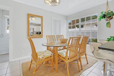 Bright coastal-style dining room with a glass round table and six rattan chairs on a jute rug, large window, hanging plant, and breakfast bar stools