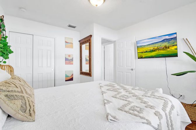 Bright modern white bedroom interior with textured bedding and patterned throw, wall-mounted TV showing green hills, wooden full-length mirror, closet doors and a potted plant.