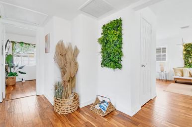 Sunlit modern hallway with warm hardwood floors, white walls, a wall-mounted vertical green plant panel, woven baskets holding dried palm fronds and a magazine, and glimpses of a cozy, plant-filled living area.