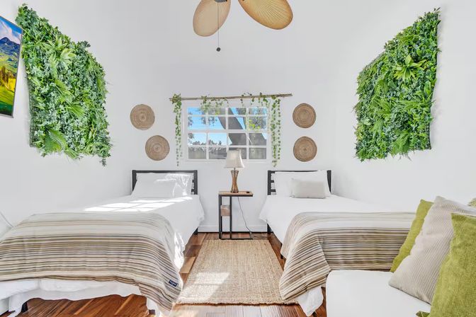 Sunny twin bedroom with two white-made beds, striped throws, central nightstand and lamp, leafy green wall panels, vine-framed window, woven wall baskets, rattan rug and hardwood floors — modern tropical vibe.