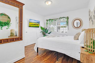 Sunny boho bedroom with white bedding, hardwood floors, rattan mirrors and lamps, lush houseplants, wall-mounted TV, and a sunlit window for a coastal cottage vibe.