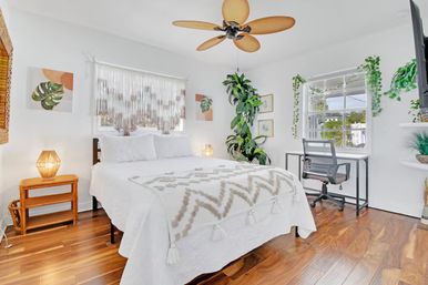 Bright boho bedroom with white bed and tasseled chevron throw, warm wood floors, rattan nightstand and lamp, large wooden-blade ceiling fan, hanging plants and a small desk by a sunlit window.