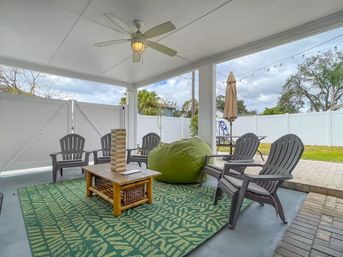 Covered residential backyard patio with ceiling fan, green patterned rug, wooden coffee table with Jenga tower, green beanbag and gray Adirondack-style chairs, string lights and white vinyl fence