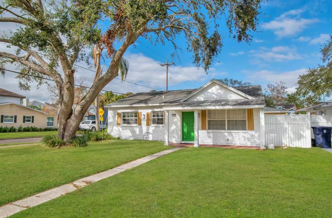Sunlit single-story white bungalow with a bright green front door, wooden shutters, large oak tree and manicured lawn on a suburban corner lot under a blue sky.