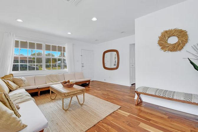 Sunny coastal-style living room with large grid window, white walls, long sectional sofa, wicker coffee table on a jute rug and warm hardwood floors with neutral decorative accents.