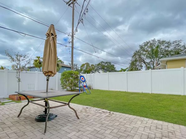 Suburban backyard with paver patio, metal table and closed beige umbrella, green lawn and white vinyl fence, a freestanding bullseye target by shrubs, and a cloudy sky with overhead utility lines — ready for backyard target practice.