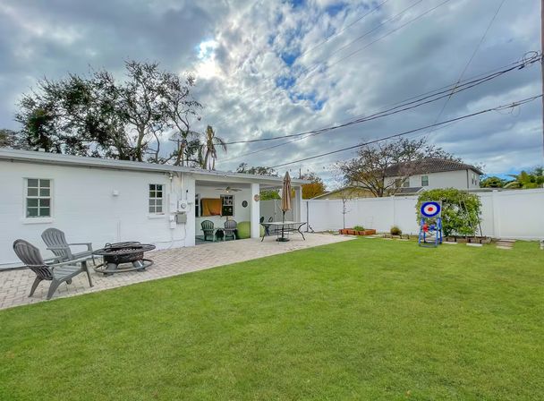 Suburban backyard with green lawn and paved patio beside a white single-story house, Adirondack chairs around a fire pit, patio table with umbrella, a target stand by a white privacy fence, and a dramatic cloudy sky.
